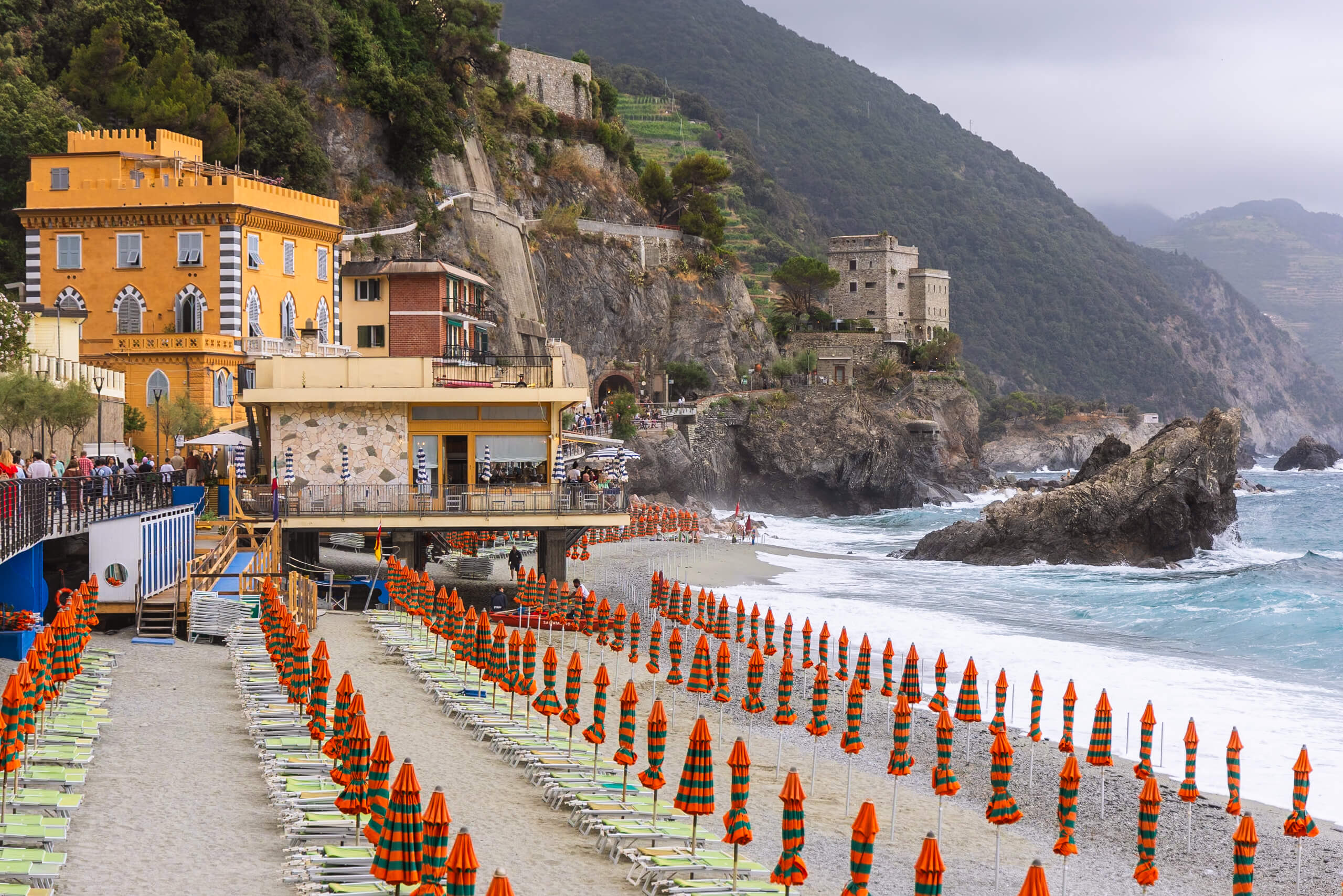 A vibrant view of the beach in Monterosso al Mare, Cinque Terre, with rows of orange and green striped umbrellas lining the sandy shore. The turquoise waters meet the mountainous coastline, creating a picturesque scene. This bustling beach is a popular spot in any Cinque Terre itinerary, especially for a one-day visit.