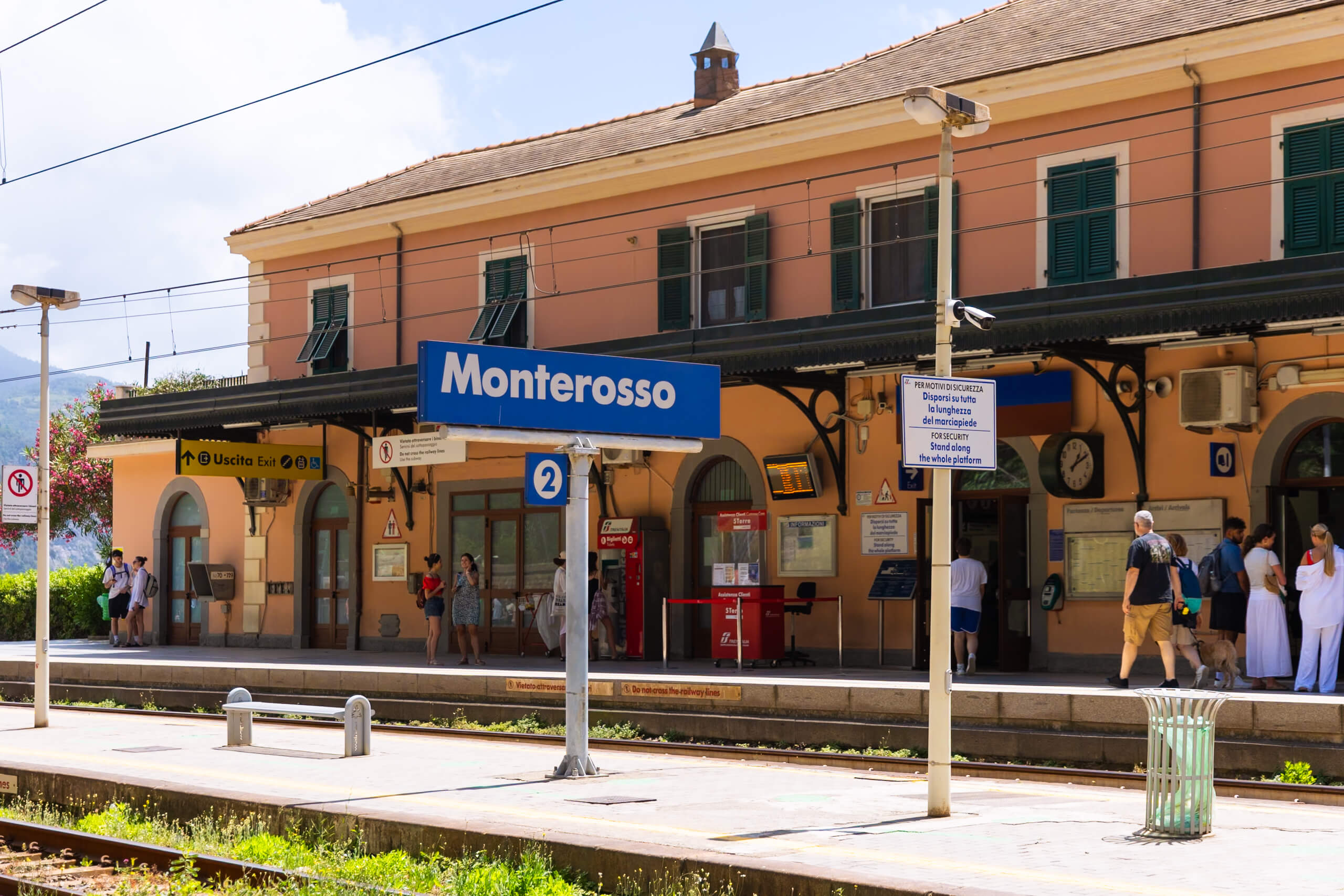The Monterosso train station in Cinque Terre, featuring a prominent blue sign and an orange-colored building. Passengers wait on the platform under a clear sky.