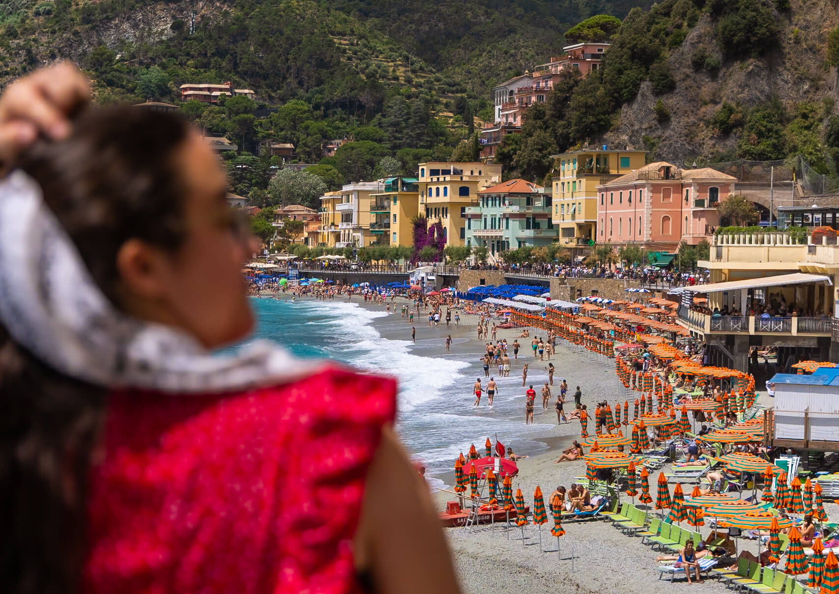 A scenic view of Monterosso al Mare beach from a viewpoint, with Penelope blurred in the foreground. The colorful beach umbrellas, busy shoreline, and lush hills create a picturesque backdrop. A great spot for capturing memories on a one-day visit to Cinque Terre.