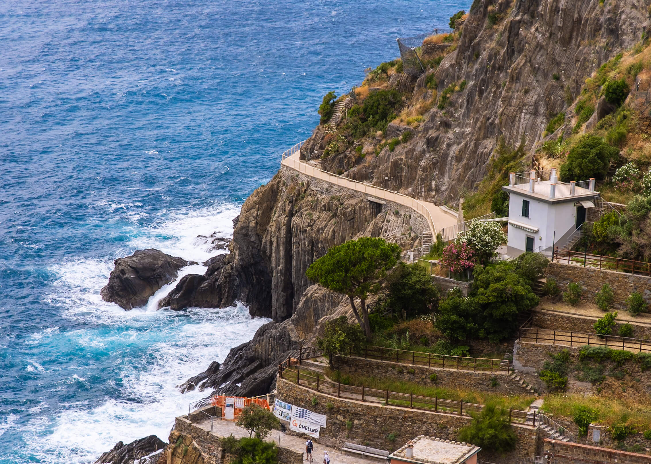 A picturesque coastal pathway winding along the cliffs of Riomaggiore, with turquoise waves crashing against the rocks below. The scene includes a small white building and terraced gardens.