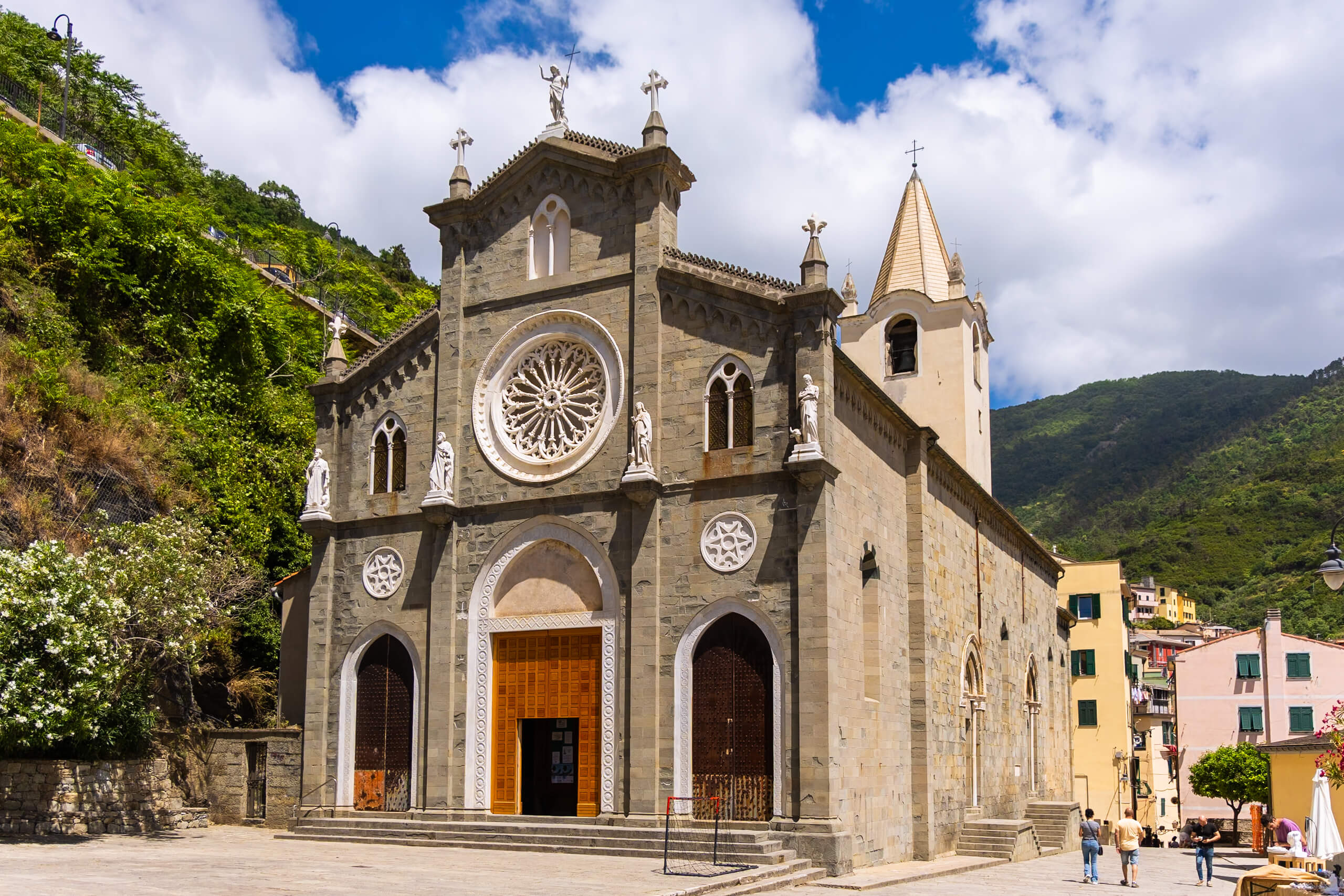 The facade of a historic church in Riomaggiore, featuring intricate stone carvings and statues. The church has a large rose window and is set against a backdrop of green hills.