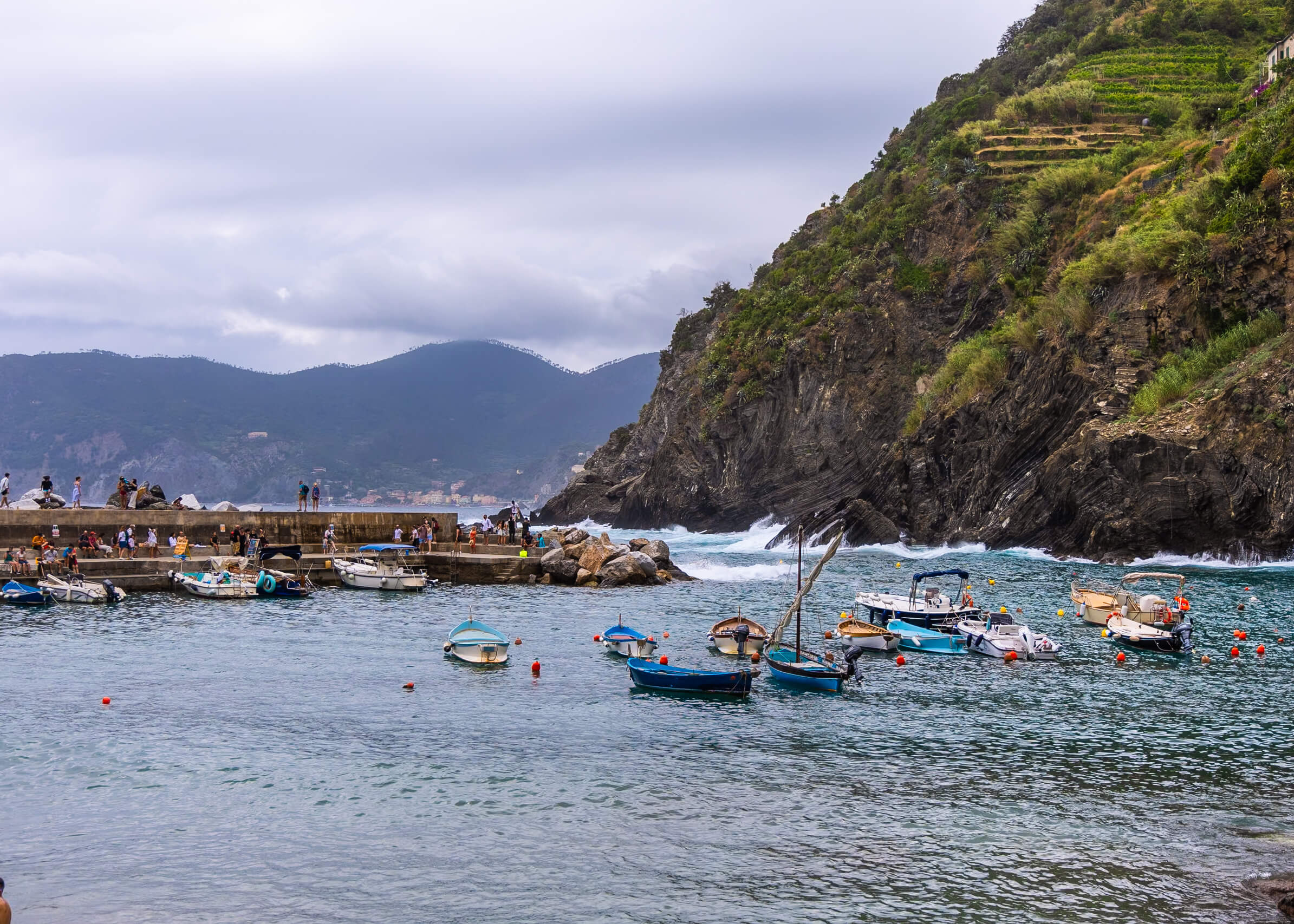 Vernazza's picturesque harbor, surrounded by rugged cliffs and small boats floating in the calm waters. The distant hills create a stunning backdrop under a cloudy sky, capturing the natural beauty of Cinque Terre. A perfect view for a one-day visit to the area.