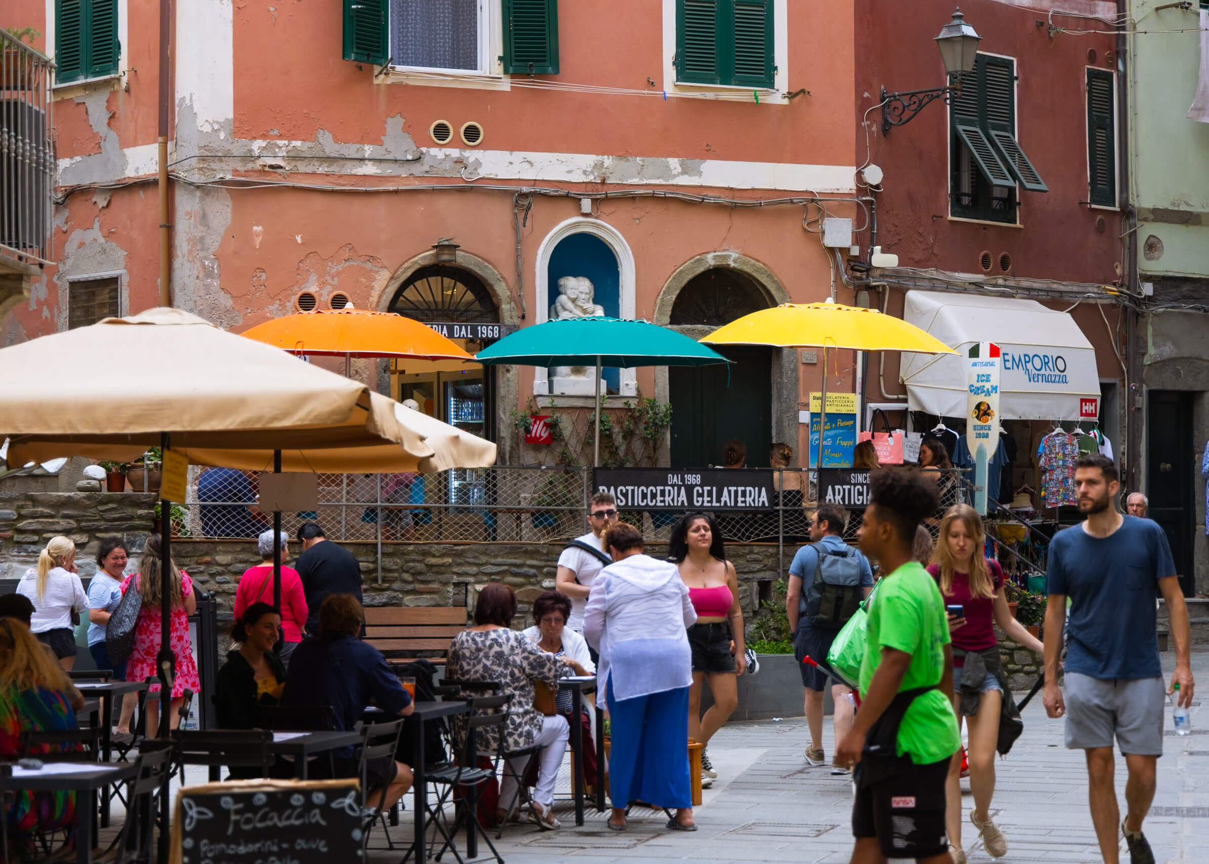 A colorful and lively street scene in Vernazza, Cinque Terre, featuring people walking past a historic building with a gelateria. The warm, inviting ambiance of the piazza makes it a great spot to relax and enjoy local treats during a one-day visit to Cinque Terre.