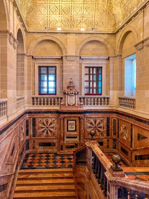 Staircase inside the Archivo de Indias with marble railings. patterned tile floors. stone arches. and a carved ceiling. A sign above the lower doorway reads "Archivo General de Indias."