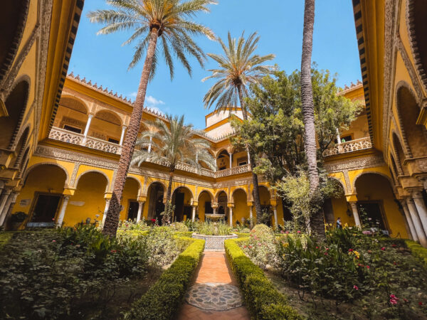 Sunny courtyard inside the Royal Alcázar of Seville with yellow arched walls. palm trees. flower beds. and a small tiled fountain. Visiting the palace gardens is one of the best things to do in Seville for people who love historic architecture and quiet green spaces.