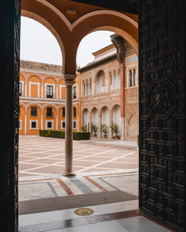 Courtyard framed by a dark wooden doorway with orange walls. white arches. brick details. and patterned tile flooring. The open space shows Seville’s mix of Moorish and Spanish architecture in a quiet historic setting.
