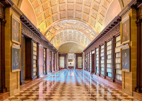 Long gallery inside the Archivo de Indias with polished checkered floors. tall archive shelves. framed portraits. and a glowing arched ceiling. Visiting this historic building is one of the best things to do in Seville for free for travelers interested in architecture and history.