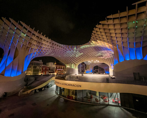 Las Setas lit up at night with blue. green. and purple lights glowing beneath the wooden lattice canopy. A sign on the lower level reads "Mercado de la Encarnacion."