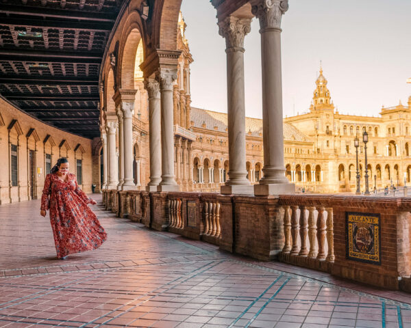 Penelope stands in a red patterned dress under the covered arcade at Plaza de España. White columns frame the glowing brick plaza in the background and a tile sign on the wall reads "Alicante Exposicion Ibero Americana."