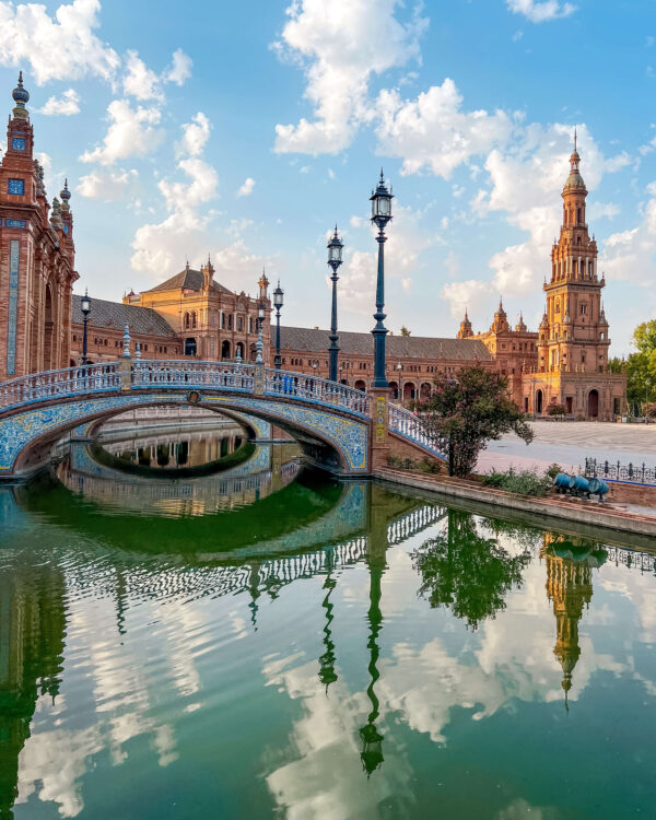 Blue and white tiled bridge at Plaza de España crossing over the green canal with towers and brick buildings reflected in the water. The open plaza and dramatic architecture make this one of the best things to do in Seville for free.