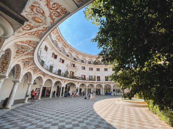 Curved courtyard at Plaza del Cabildo with white arcades. painted arch details. a black and white patterned stone floor. and a large leafy tree casting shade. This peaceful hidden plaza is one of the best things to do in Seville for free.