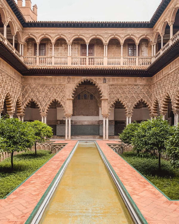 Courtyard inside the Real Alcázar of Seville with a long rectangular pool. orange tiled walkways. trimmed greenery. and intricate scalloped arches. The detailed carvings and quiet symmetry make the palace one of the best things to do in Seville.
