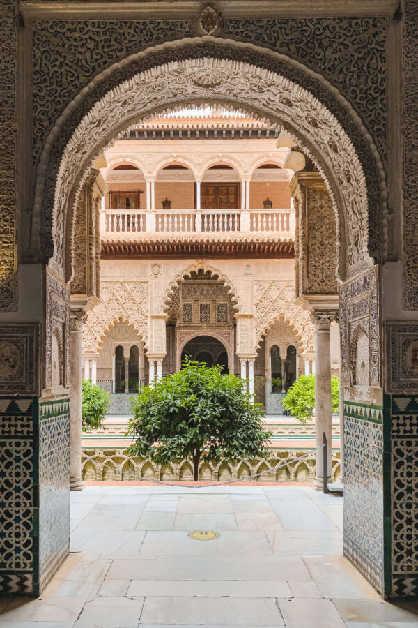 Ornate archway inside the Real Alcázar of Seville framing a courtyard filled with scalloped arches. carved plaster walls. geometric tilework. and leafy orange trees. The layered view highlights the palace’s Islamic and Mudéjar design details.
