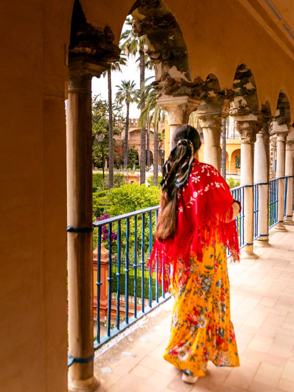 Penelope stands on an upper arcade inside the Real Alcázar wearing a yellow floral dress and a red embroidered shawl with fringe. Weathered columns. rounded arches. blue railings. and palm filled gardens surround the walkway.