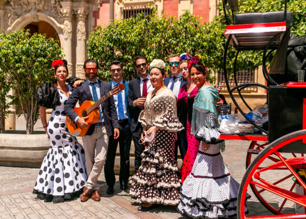 Group of people dressed for Feria de Abril posing beside a bright red horse carriage in Seville. Several women wear polka dot flamenco dresses and shawls while one man holds a guitar in the sunny courtyard.