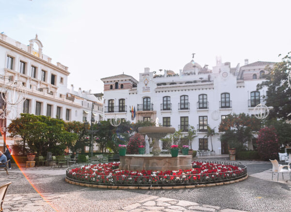 A flower lined fountain sits in a sunny pedestrian square surrounded by white buildings and outdoor seating in Estepona. A building behind the fountain displays the words "Hotel El Pilar Andalucia Hotel Boutique".