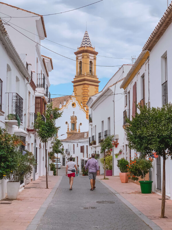 A quiet Estepona street leads toward a white church with a tall yellow bell tower and patterned spire. Whitewashed homes with balconies trees and colorful flower pots line both sides of the road.
